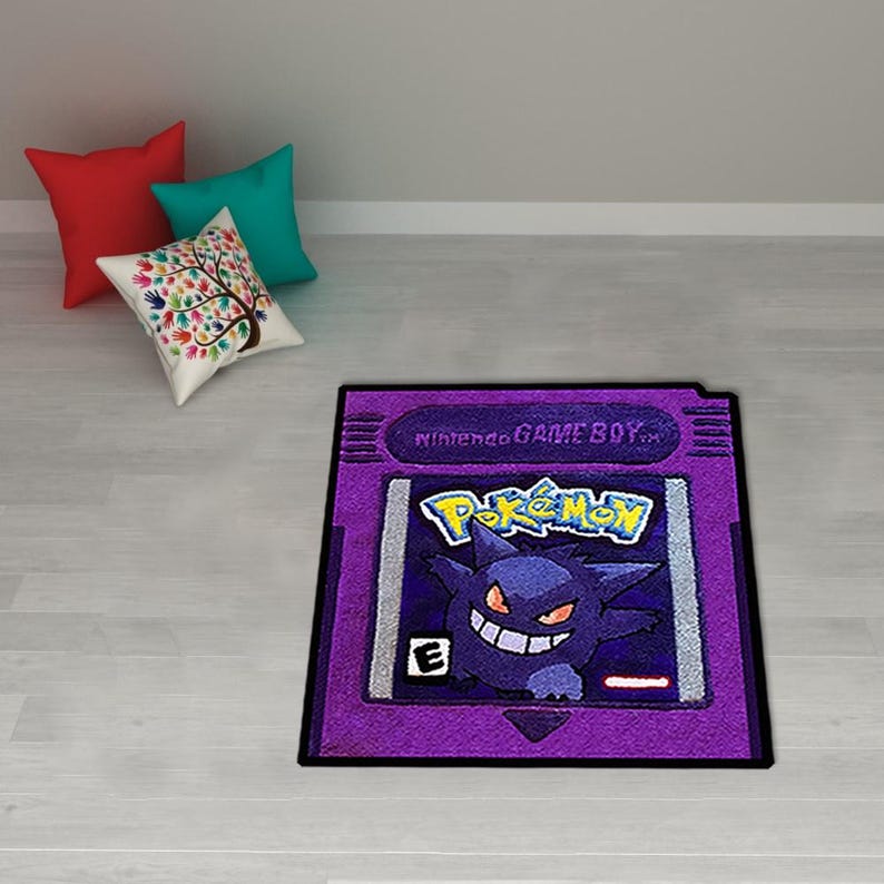 May include: A purple rug shaped like a Nintendo Game Boy cartridge, featuring the Pok&eacute;mon logo and a Gengar character. The rug has a black border and is placed on a light-colored floor. Three throw pillows in red, teal, and white with a tree design are in the background.