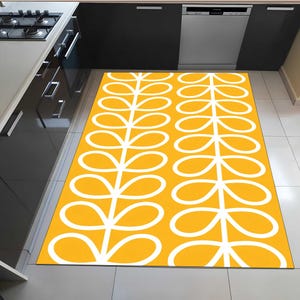 May include: A rectangular, yellow kitchen rug with a white, stylized leaf pattern. The rug is placed on a tiled floor in front of a dishwasher and cabinets. The design is a repeating pattern of connected, oval shapes.