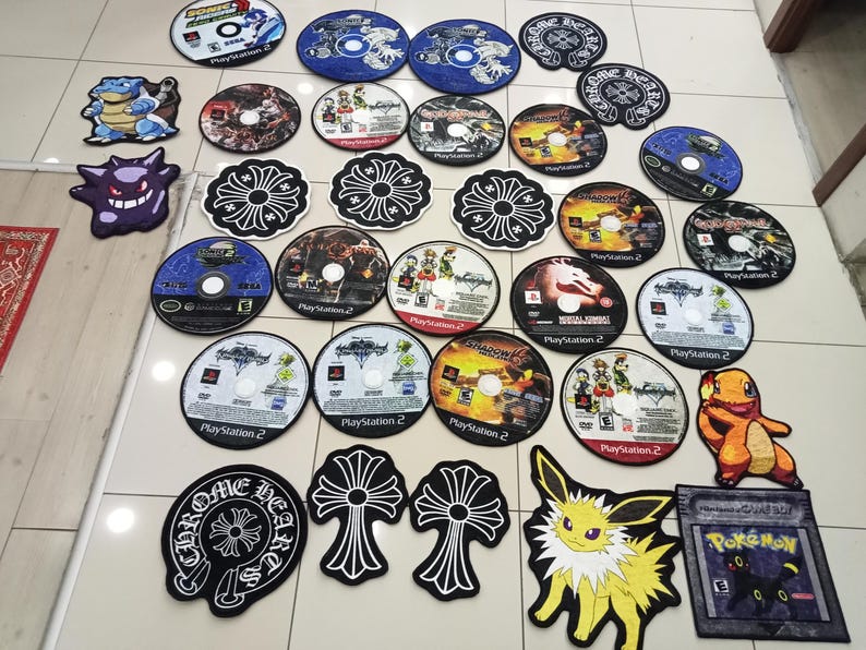 May include: A collection of various round and shaped rugs. The rugs feature designs of video game discs, cartoon characters, and a floral pattern. The colors include blue, black, white, orange, and yellow. The rugs are made of a soft material.