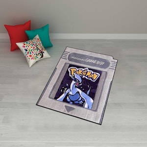 May include: A rectangular rug designed to resemble a Nintendo Game Boy cartridge. The rug features the "Pokemon" logo and a blue and white creature against a dark background. The rug is gray with a white border.