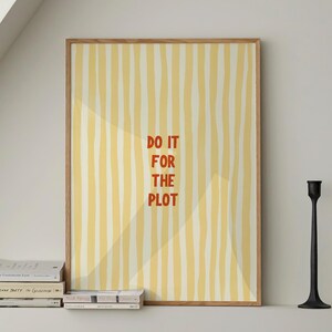 Puede incluir: Un póster enmarcado con un patrón de rayas amarillas y blancas y el texto "DO IT FOR THE PLOT" en rojo. El póster se exhibe encima de una estantería con libros y dos candelabros negros.