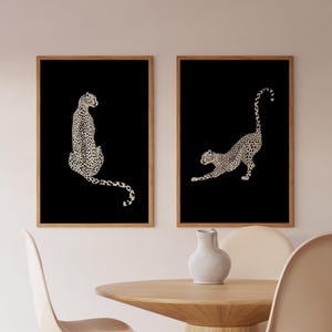 May include: Two framed art prints featuring cheetah illustrations. The cheetahs are depicted in a cream color with black spots against a black background. The frames are a light brown color. The prints are displayed in a dining room setting.