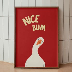 May include: A framed art print with a red background and the words "NICE BUM" in yellow. A white goose with an orange beak and a curved neck is featured below the text. The frame is brown.