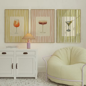 May include: Three framed art prints with cocktail illustrations. Each print shows a different cocktail in a glass, set against a striped background. The frames are gold-coloured. The prints are above a white cabinet and a yellow chair.