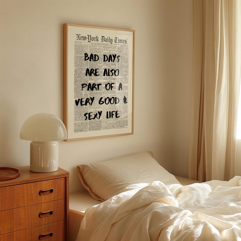 May include: A framed print of a newspaper page with the headline "New-York Daily Times." Bold black text reads "BAD DAYS ARE ALSO PART OF A VERY GOOD SEXY LIFE." The print hangs above a bed with white linens and a wooden nightstand.