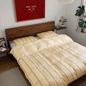 May include: A bed with a wooden headboard featuring a pale yellow duvet cover and two matching pillows, all decorated with thin vertical brown stripes. Above the bed hangs a large rectangular red framed print with the text HOW LUCKY ARE WE.