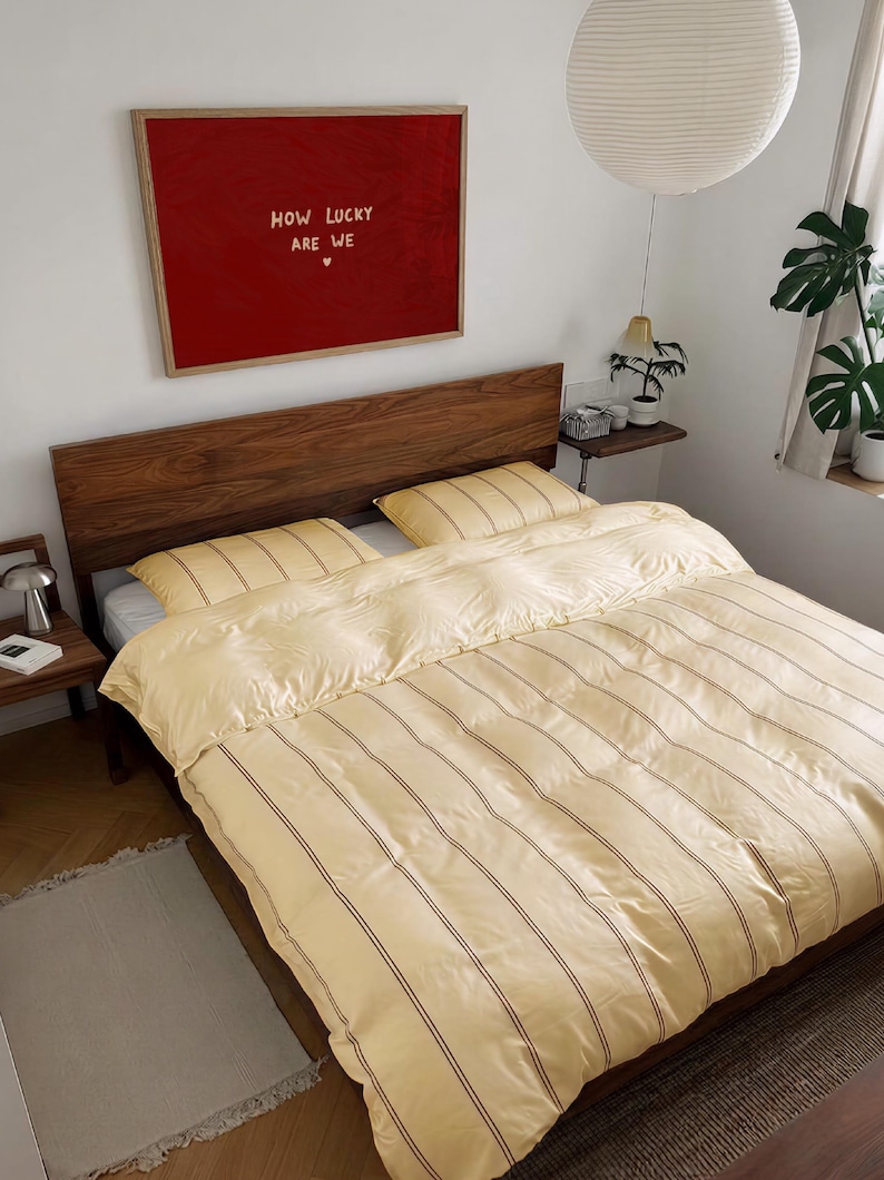 May include: A bed with a wooden headboard featuring a pale yellow duvet cover and two matching pillows, all decorated with thin vertical brown stripes. Above the bed hangs a large rectangular red framed print with the text HOW LUCKY ARE WE.