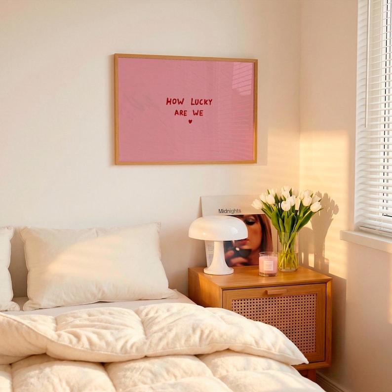 May include: A rectangular wall art print featuring a solid pink background with the red text "HOW LUCKY ARE WE" centered, followed by a small red heart symbol. The print is enclosed in a thin, light-toned wooden frame and mounted on a wall.