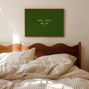 May include: A rectangular wall art piece in a thin wooden frame featuring a dark green background with the text "HOW LUCKY ARE WE" in a light cream color, positioned above a small heart symbol, centered on the wall above a wooden headboard.