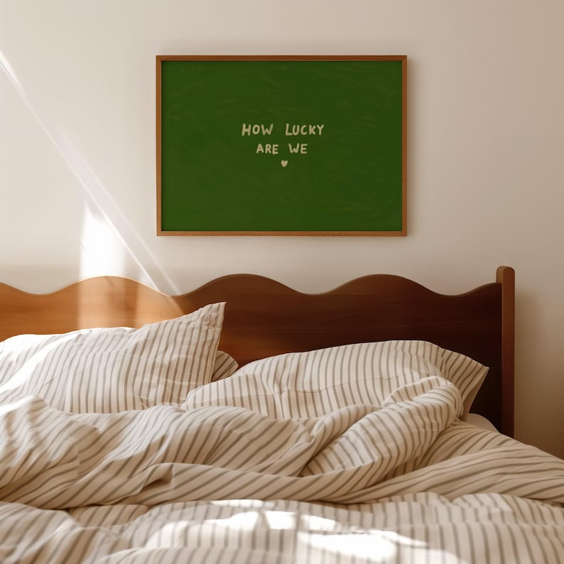 May include: A rectangular wall art piece in a thin wooden frame featuring a dark green background with the text "HOW LUCKY ARE WE" in a light cream color, positioned above a small heart symbol, centered on the wall above a wooden headboard.