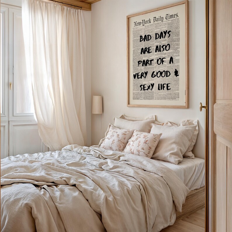 May include: A bedroom scene featuring a bed with beige bedding and floral pillows. A framed print on the wall reads "BAD DAYS ARE ALSO PART OF A VERY GOOD SEXY LIFE." Soft, sheer curtains frame a window.