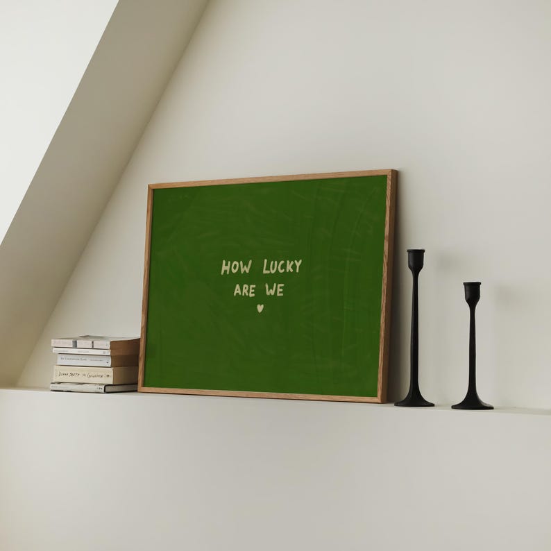 May include: A rectangular wall art print in a light wood frame featuring a deep green background with the text "HOW LUCKY ARE WE" in a light cream color, centered above a small heart symbol, displayed alongside a stack of books and two black candlesticks.