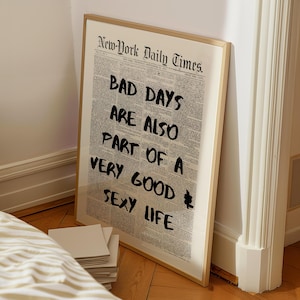 May include: A framed print featuring a vintage newspaper background with the headline "New-York Daily Times." Bold black text reads "BAD DAYS ARE ALSO PART OF A VERY GOOD SEXY LIFE." The frame is a light wood color, and the print leans against a white wall.