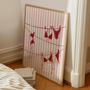May include: Framed art print featuring a red and white striped background with red bikini illustrations hanging on clotheslines. The artwork is displayed in a light wood frame, leaning against a wall on a wooden floor.