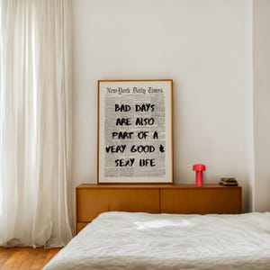May include: A framed print with the text "BAD DAYS ARE ALSO PART OF A VERY GOOD SEXY LIFE" in black font. The print is on a newspaper-style background and is displayed above a wooden cabinet. A red lamp and books are on the cabinet.