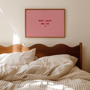 May include: A rectangular wall art print in a wooden frame featuring a solid pink background with the red text "HOW LUCKY ARE WE" centered, followed by a small red heart symbol. The print is displayed on a plain wall above a wooden headboard.