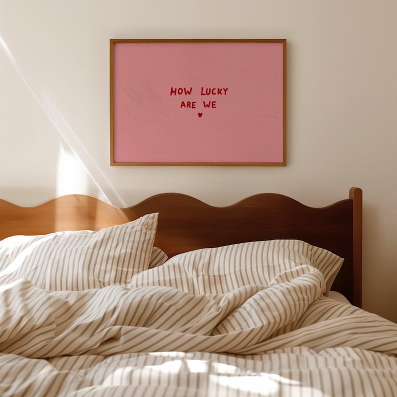 May include: A rectangular wall art print in a wooden frame featuring a solid pink background with the red text "HOW LUCKY ARE WE" centered, followed by a small red heart symbol. The print is displayed on a plain wall above a wooden headboard.