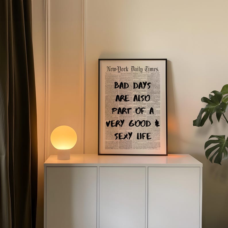 May include: A framed print with the text "BAD DAYS ARE ALSO PART OF A VERY GOOD SEXY LIFE" in black against a newspaper background. A white cabinet with a round, glowing lamp sits below the print.