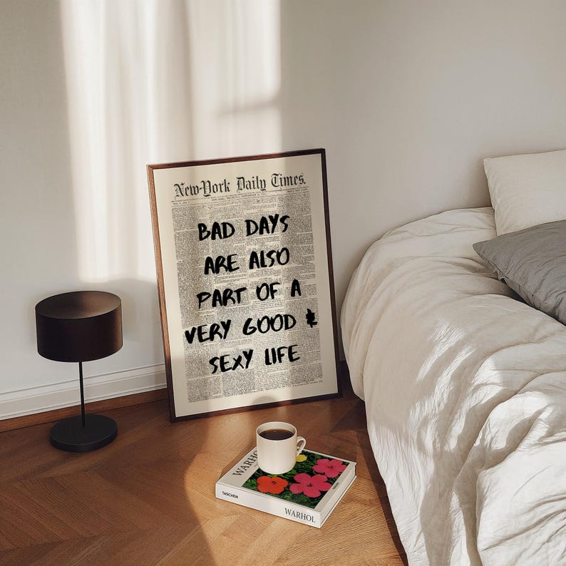 May include: A framed print with the text "BAD DAYS ARE ALSO PART OF A VERY GOOD SEXY LIFE" in a newspaper-style layout. A coffee cup sits on a book with floral art. A black lamp and a bed are also in the image.