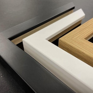 May include: Three L-shaped corner sections of picture frames arranged in a nested stack. The outermost frame is matte black, the middle frame is solid white, and the innermost frame features a natural light brown wood grain finish.