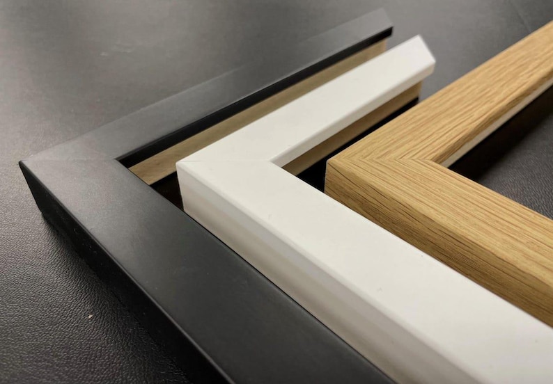 May include: Three L-shaped corner sections of picture frames arranged in a nested stack. The outermost frame is matte black, the middle frame is solid white, and the innermost frame features a natural light brown wood grain finish.