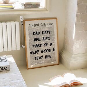 May include: A framed print with the text "BAD DAYS ARE ALSO PART OF A VERY GOOD SEXY LIFE" in black over a newspaper background. The frame is light brown. The print is leaning against a wall near a window.