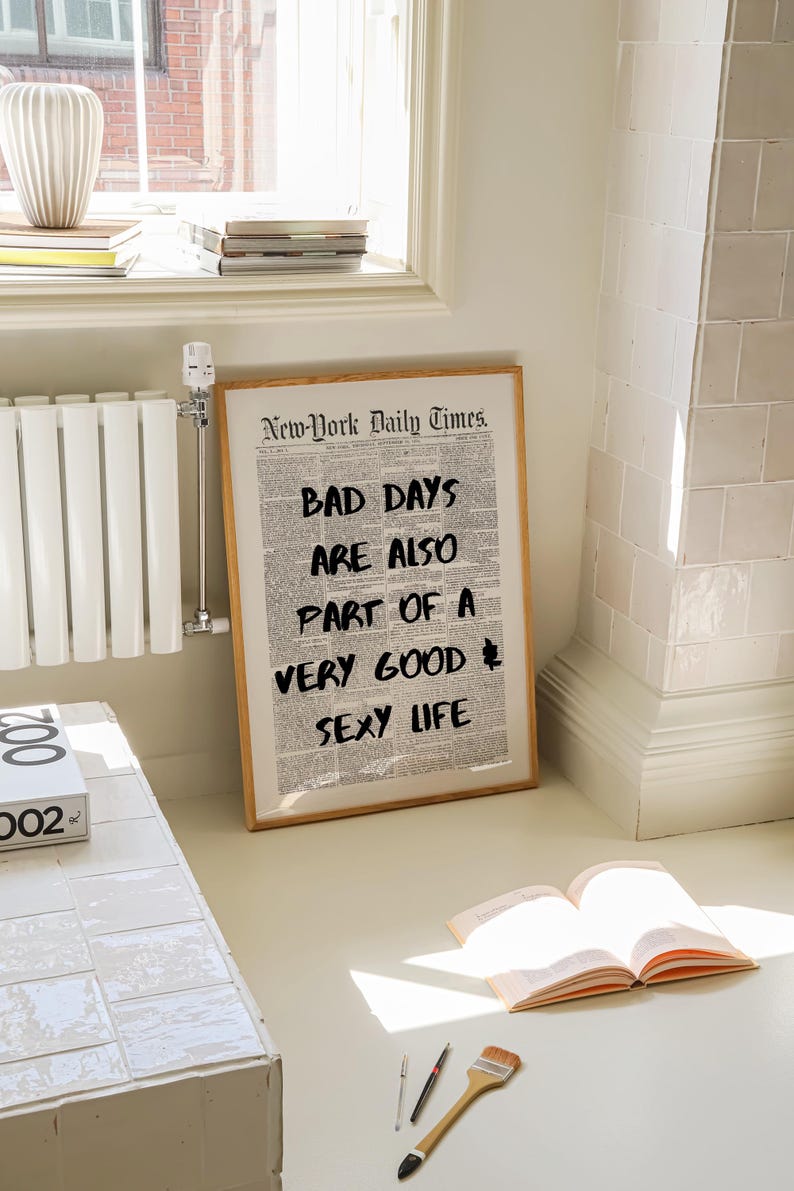 May include: A framed print with the text "BAD DAYS ARE ALSO PART OF A VERY GOOD SEXY LIFE" in black over a newspaper background. The frame is light brown. The print is leaning against a wall near a window.