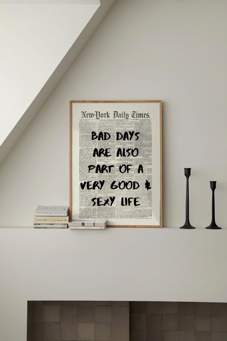 May include: A framed print featuring a newspaper background with the text "BAD DAYS ARE ALSO PART OF A VERY GOOD SEXY LIFE." The print is in a wooden frame, with two black candle holders to the right and a stack of books below.