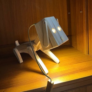 Puede incluir: Una lámpara de madera con forma de perro con un acabado marrón claro. La lámpara tiene dos agujeros redondos para los ojos y un cable con un enchufe.