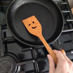 May include: A black cast iron frying pan with a wooden spatula. The spatula has a winking face carved into it. The pan is on a black hob.