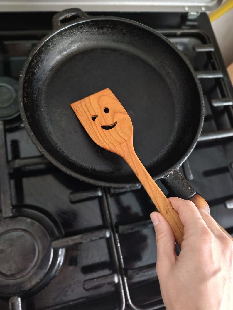May include: A black cast iron frying pan with a wooden spatula. The spatula has a winking face carved into it. The pan is on a black hob.