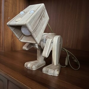 Puede incluir: Una lámpara de madera con forma de robot con una bombilla en la cabeza. La lámpara está hecha de madera marrón claro y tiene un cable.