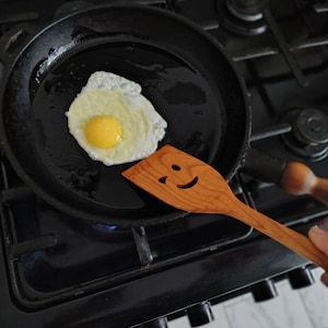 May include: A fried egg in a black frying pan with a wooden spatula. The spatula has a smiling face carved into it. The egg yolk is yellow, and the whites are cooked. The pan is on a gas hob.