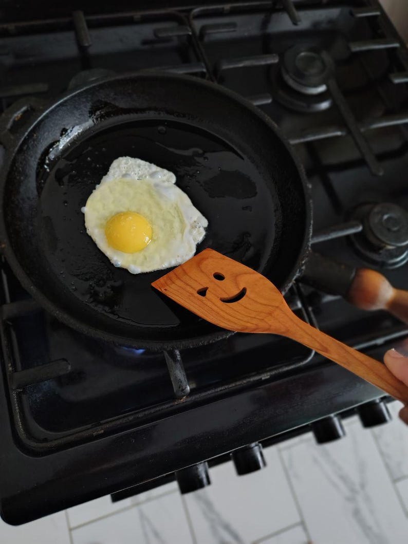 May include: A fried egg in a black frying pan with a wooden spatula. The spatula has a smiling face carved into it. The egg yolk is yellow, and the whites are cooked. The pan is on a gas hob.