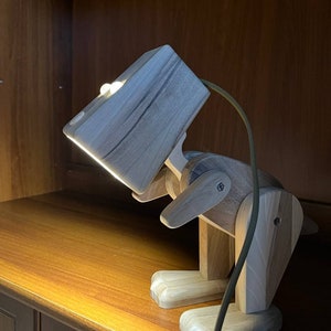 Puede incluir: Una lámpara de madera con forma de dinosaurio con una luz en la cabeza. La lámpara está en un estante de madera y está enchufada.