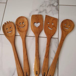 May include: Five wooden kitchen utensils with smiley faces carved into them. The utensils include a slotted spoon, a solid spoon, a spatula with a heart cutout, a spatula with a slanted edge, and a spoon. Each utensil is made of light brown wood.