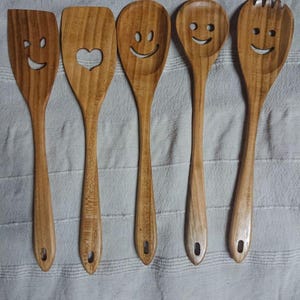 May include: A set of five wooden kitchen utensils, each with a smiling face design. The set includes a spatula, a spoon with a heart cutout, and three spoons with different smiley face designs. The utensils are made of light brown wood.