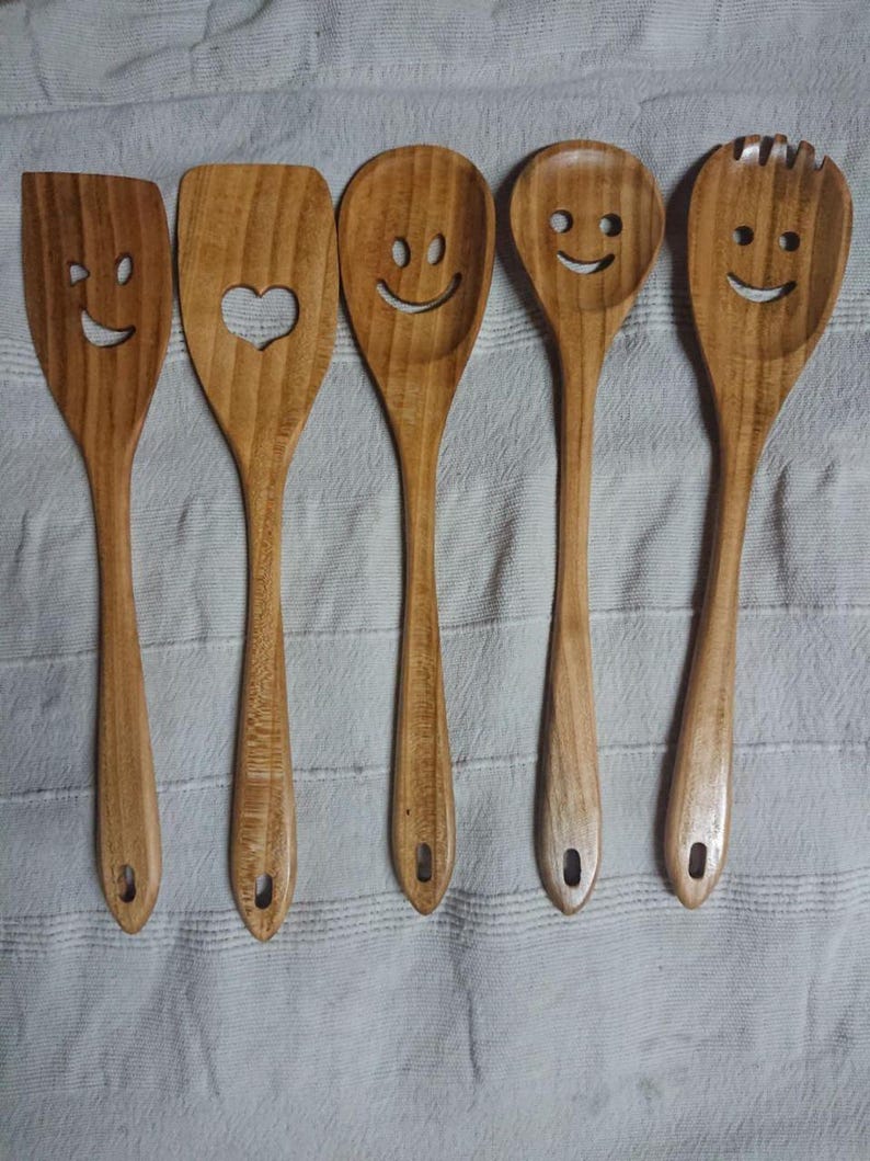 May include: A set of five wooden kitchen utensils, each with a smiling face design. The set includes a spatula, a spoon with a heart cutout, and three spoons with different smiley face designs. The utensils are made of light brown wood.