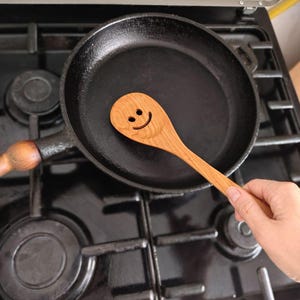 May include: A wooden spoon with a smiling face lies in a black cast iron frying pan on a gas hob. The spoon is light brown, and the pan is dark. The hob has black burners and a metal surface.