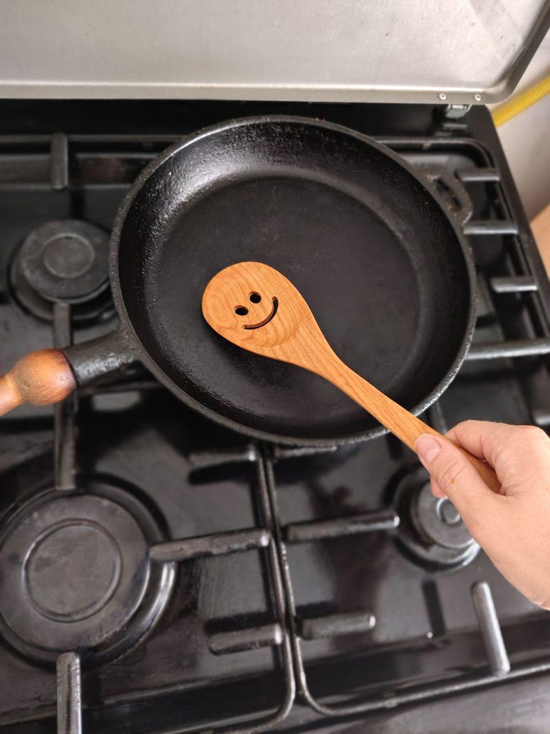 May include: A wooden spoon with a smiling face lies in a black cast iron frying pan on a gas hob. The spoon is light brown, and the pan is dark. The hob has black burners and a metal surface.