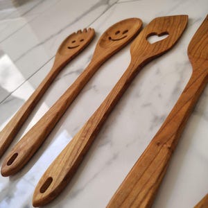 May include: A selection of wooden kitchen utensils, including a slotted spoon with a heart-shaped cutout, a fork-like spoon, and spoons with smiley faces. The utensils are made of light brown wood and have a smooth finish. Each utensil has a hole in the handle.