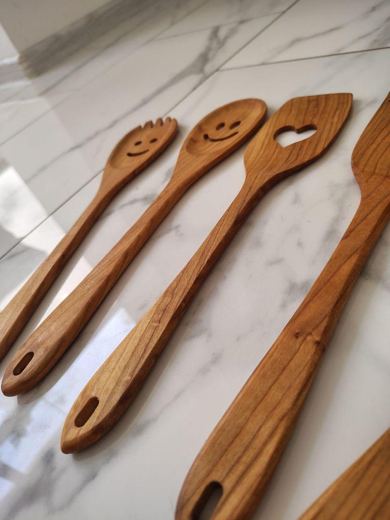 May include: A selection of wooden kitchen utensils, including a slotted spoon with a heart-shaped cutout, a fork-like spoon, and spoons with smiley faces. The utensils are made of light brown wood and have a smooth finish. Each utensil has a hole in the handle.