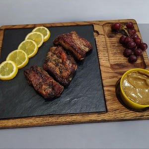 May include: Wooden serving board with a black slate insert, holding grilled ribs, lemon slices, grapes, and a small yellow bowl of sauce. The board has a natural wood grain finish.