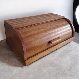 Puede incluir: Una panera de madera con tapa enrollable y diseño curvo. La caja es de madera marrón claro y tiene un asa en la parte delantera. La parte superior de la caja tiene una textura acanalada.