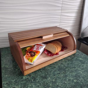 May include: A wooden bread box with a roll-top lid. Inside, there's a loaf of sliced bread on a red and white patterned cloth and a package of bread. The box is made of light brown wood and sits on a green countertop.