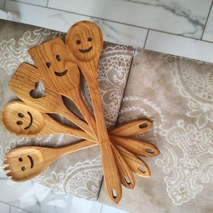May include: A selection of wooden kitchen utensils, including spoons, a spatula with a heart cutout, and a fork. Each utensil has a carved smiley face. The wood has a warm, natural tone.