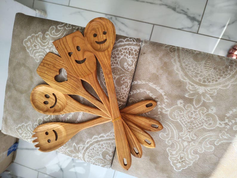 May include: A selection of wooden kitchen utensils, including spoons, a spatula with a heart cutout, and a fork. Each utensil has a carved smiley face. The wood has a warm, natural tone.
