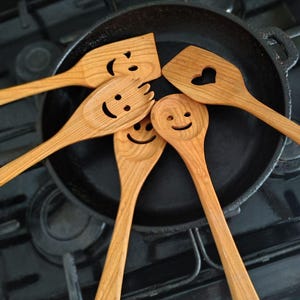 May include: A selection of wooden kitchen utensils, including spatulas and spoons, arranged around a black cast iron skillet. The utensils feature carved smiley faces and a heart-shaped cutout, adding a playful touch to the cooking tools.