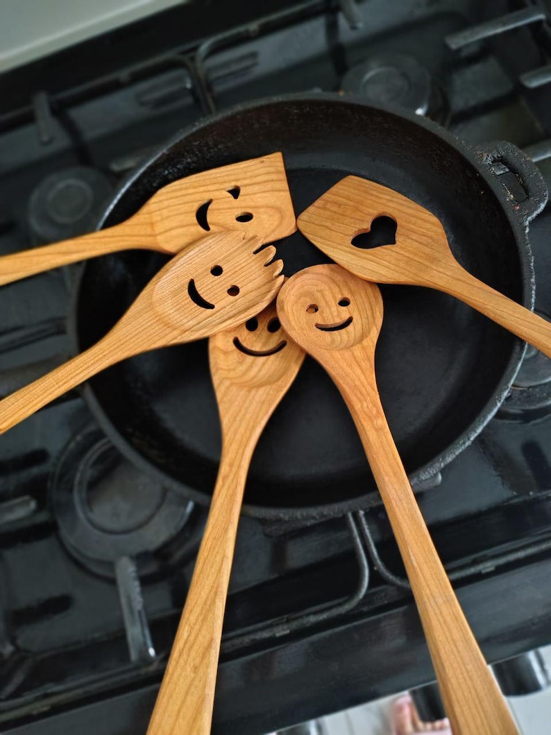 May include: A selection of wooden kitchen utensils, including spatulas and spoons, arranged around a black cast iron skillet. The utensils feature carved smiley faces and a heart-shaped cutout, adding a playful touch to the cooking tools.