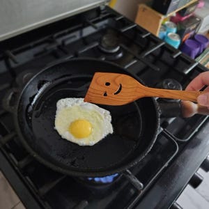 May include: A fried egg in a black frying pan on a gas hob. A wooden spatula with a smiley face is used to flip the egg. The egg has a yellow yolk and white egg whites. The hob is black.